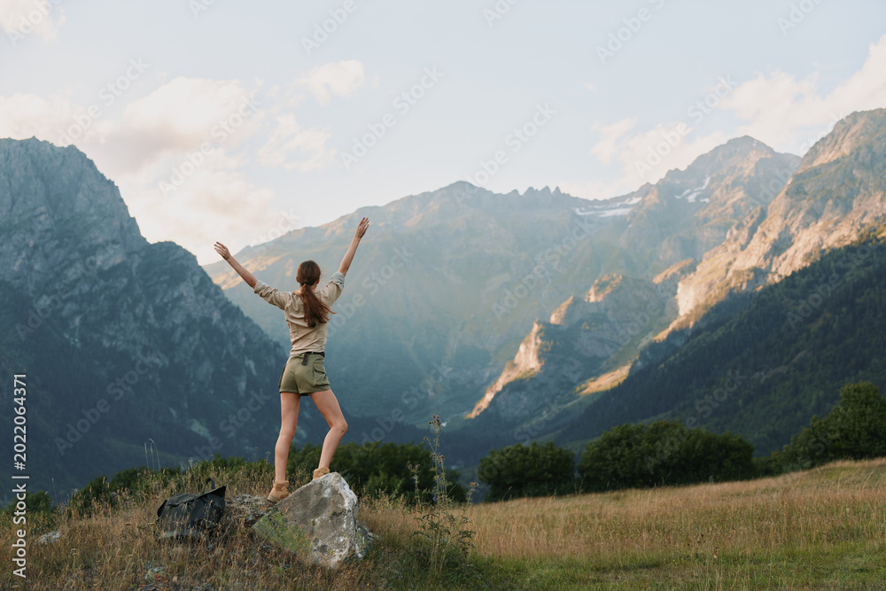 Fototapeta premium Young woman outdoors lifestyle on a rock with arms raised in a scenic mountain landscape, celebrating nature, hiking, freedom, and adventure under bright daylight.