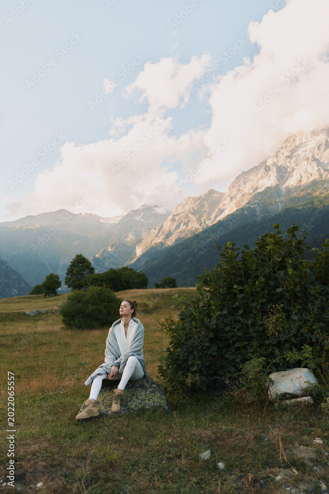 Fototapeta premium Young woman in a light denim jacket and jeans sits on a rock in a green meadow, with distant alpine mountains and a bright sky forming a dramatic natural backdrop