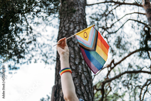 Hand raises progress pride flag next to tree trunk in forest. Human rights activism, support for LGBTQ community, non-binary person lifestyle.