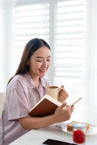 Smiling young asian woman having breakfast, coffee and bread  while enjoy reading book . morning routine.