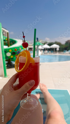 Hand holding a colorful cocktail with orange slice and cherry, poolside view featuring water slide and sun loungers in a resort setting