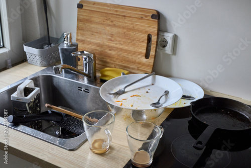 Billede på lærred A messy kitchen sink counter filled with dirty dishes, unwashed glass mugs, greasy plates with cutlery, and a cutting board