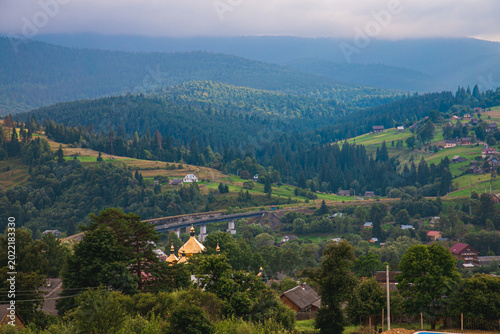 Picturesque mountain landscape. Ukrainian Carpathians. A cozy village lies in the valley below, surrounded by rolling green hills and misty mountain peaks. Bright traditional houses and untouched