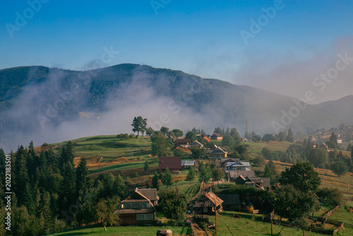 Picturesque mountain landscape. Ukrainian Carpathians. A cozy village lies in the valley below, surrounded by rolling green hills and misty mountain peaks. Bright traditional houses and untouched