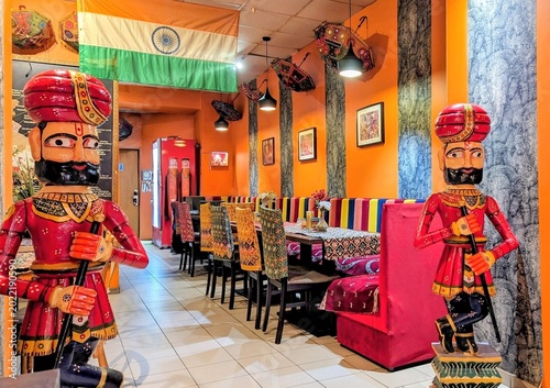 Colorful Indian restaurant interior with traditional statues, Indian flag, bright orange walls, patterned fabrics, decorative umbrellas, and vibrant dining area.