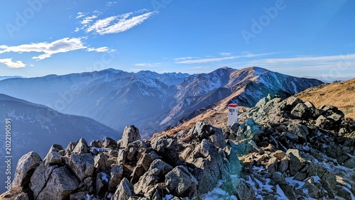 Panoramic view from a rocky mountain summit with snow patches, hiking trail marker, distant ridges, and clear blue sky.