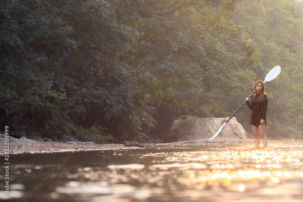 Naklejka premium Peaceful woman at river with paddle enjoying nature outdoor standing in water near forest on adventure feeling calm and confident during golden sunlight
