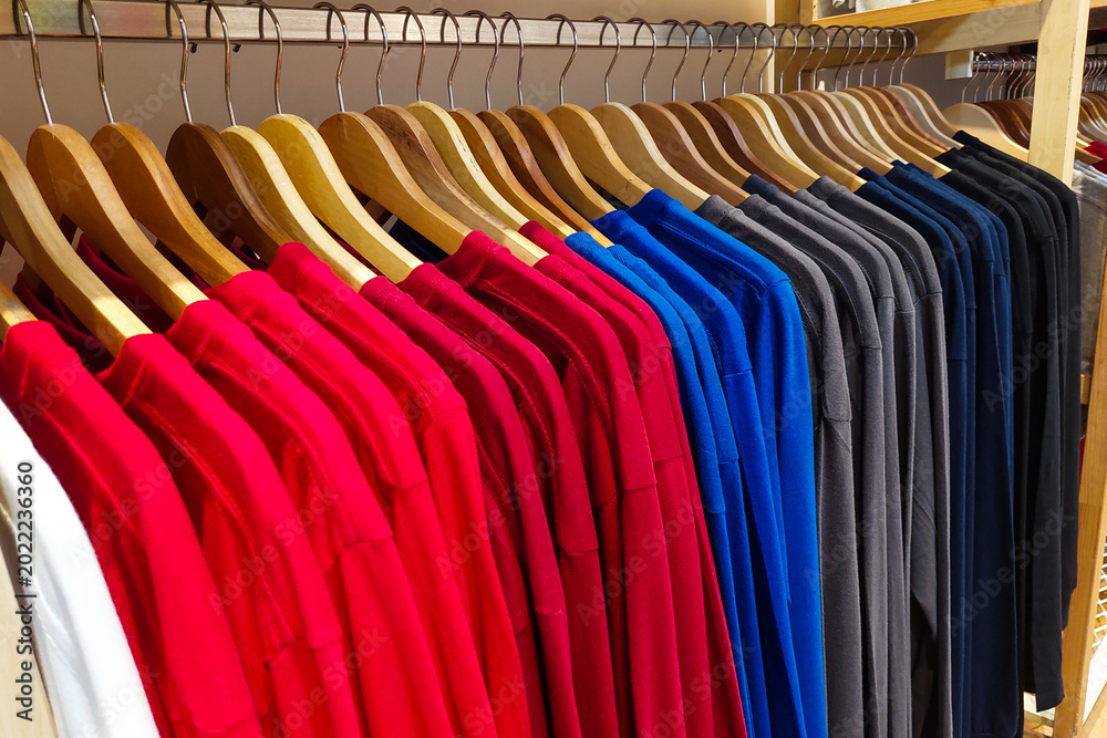 Fototapeta premium Neat rows of colorful shirts hang on wooden hangers. Bright reds, blues, and grays line a wooden clothing rack.