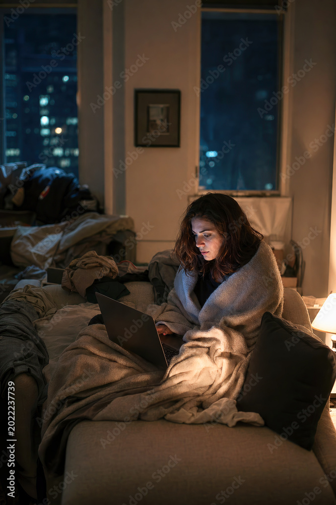 Fototapeta premium Woman working on a laptop under a blanket in a cozy apartment at night.