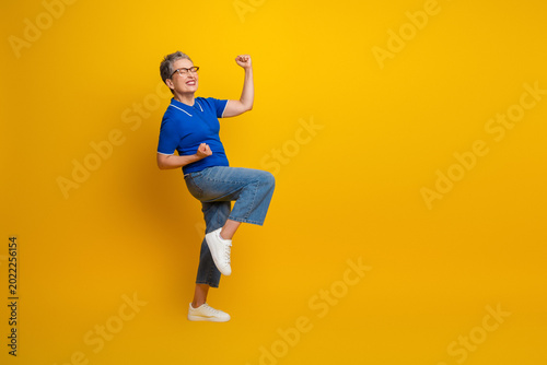 Quadro su tela Energetic senior woman jumps in blue polo shirt against yellow background expres