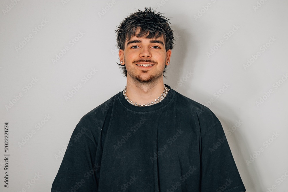 Obraz premium Young man with dark textured hair and a silver chain necklace smiling at the camera, wearing a black t-shirt against a gray studio background