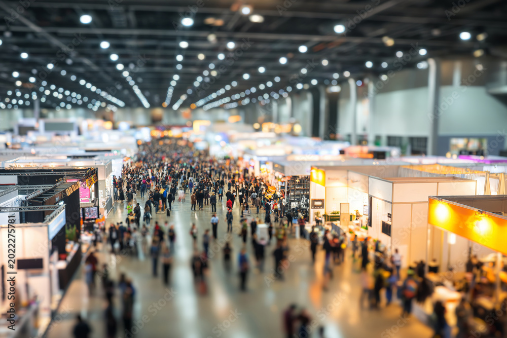 Obraz premium Blurred crowd walking through a busy trade fair exhibition hall with booths and displays, representing international business convention, product showcases, networking, and dynamic event atmosphere.