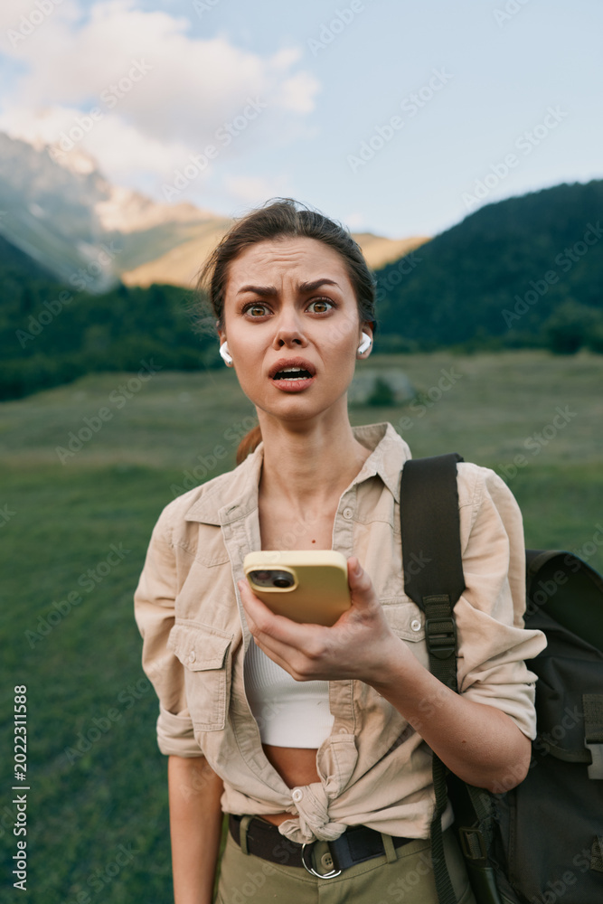 Fototapeta premium Woman traveler outdoors with backpack holds smartphone, surprised expression, mountain meadow backdrop, casual rugged style, candid lifestyle moment