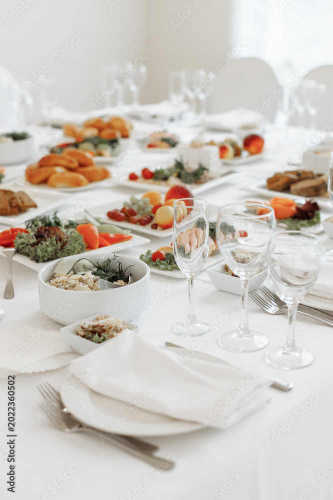 Fototapeta premium Vertical perspective looking down a long, elegantly served banquet table filled with glassware, white plates, and festive cold snacks.