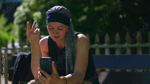 Valokuva Woman undergoing chemotherapy sitting on park bench during video call, listening