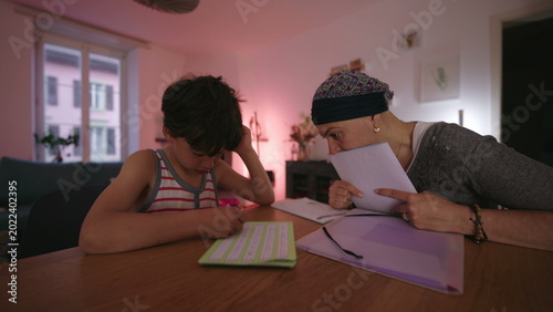 Mother undergoing chemotherapy helping child with homework at home, headscarf parent guiding learning at table, homeschooling moment showing care, patience and resilience