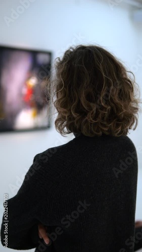 Pensive woman with curly hair viewing an abstract painting in a contemporary art gallery. The visitor, seen from behind, is contemplating the artwork hanging on a pristine white wall