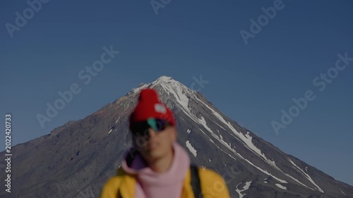 Blurry portrait of young man wearing sunglasses and knitted hat against backdrop of majestic volcano with snow-capped peak.