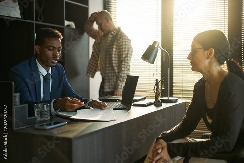 Fotografi Medium shot of Black male lawyer reviewing divorce papers with middle aged woman while Caucasian man waiting behind, representing legal separation and custody dispute