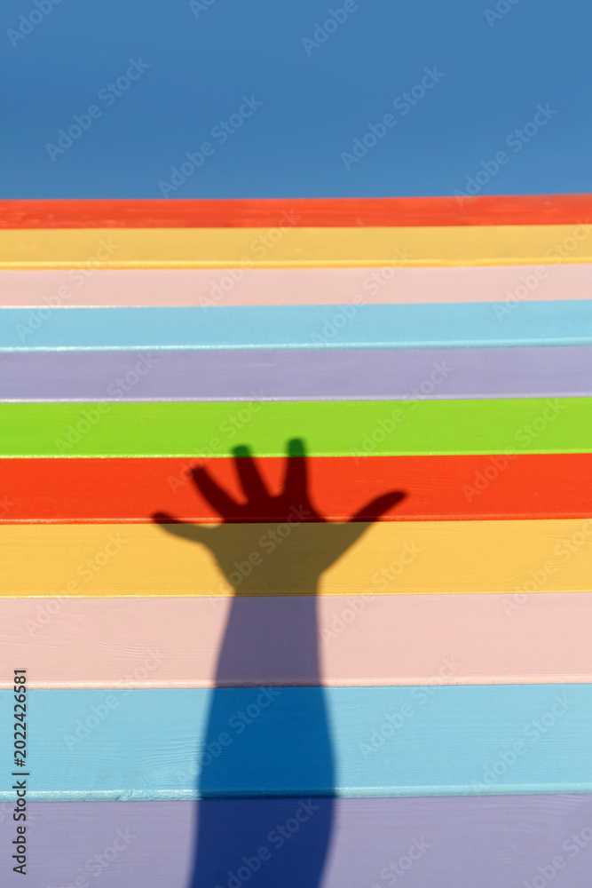 Obraz premium shadow on hand against colorful wooden fence. hand shadow, silhouette of palm on rainbow color striped background. conceptual minimal style photo representing fun, happiness, childhood, friendship