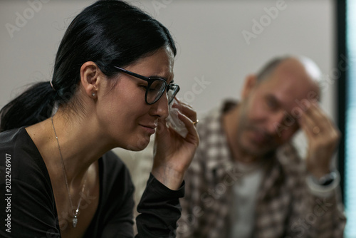 Φωτογραφία Close up of young adult woman crying and wiping tears while middle aged man sitting distressed in background, conveying divorce conflict