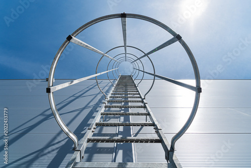 Εκτύπωση καμβά Low-angle view of a metal caged ladder attached to an industrial building facade, leading to roof access against a clear blue sky and bright sunlight, emphasizing safety, height and geometry