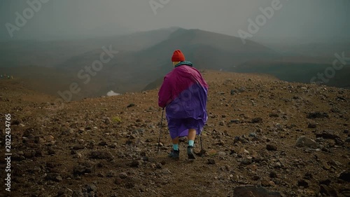 Young man in raincoat and in buff goes with trekking poles along rocky ridge high in mountains. Back view. Slow motion.
