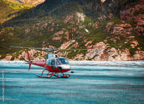 Red and silver touring helicopter landed on the textured surface of a mountain glacier with steep rocky slopes and green forests in the background.