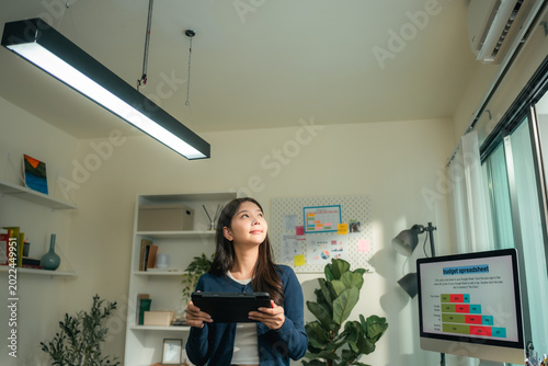 Modern woman adjusts smart lighting in her connected home office using a digital tablet, blending smart home tech, productivity, and comfort in a flexible, energy-efficient remote work environment