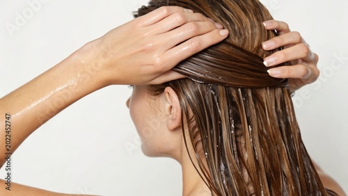 A woman with long brown hair applies conditioner or hair treatment, hands massaging and smoothing the product through her wet hair from the back.