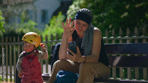 Mother undergoing chemotherapy sitting on park bench waving during video call on smartphone, smiling while toddler hands back tangerine before walking off to play, candid family moment