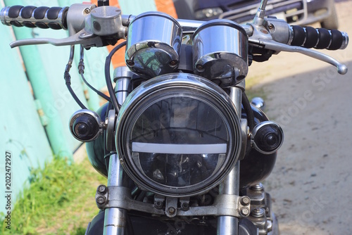 part of an old motorcycle with a white glass headlight and black handlebars near a gas tank on a summer street