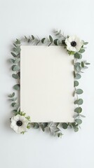 Blank paper card surrounded by eucalyptus branches and white anemone flowers on neutral background for wedding invitations and stationery design.