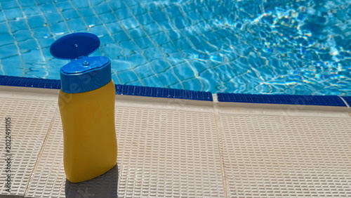 Yellow sunscreen bottle with blue cap placed on textured poolside near sparkling blue water of swimming pool under bright sunlight