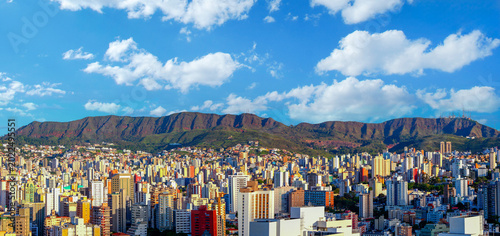 Panoramic view of Belo Horizonte skyline with the iconic Serra do Curral mountains under a blue cloudy sky. Urban landscape featuring dense high-rise buildings and natural mountain preservation.