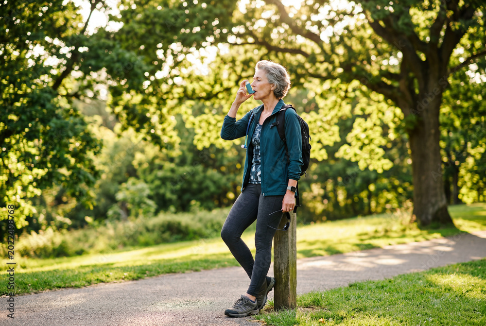 Obraz premium Mature woman with backpack standing outdoors using an asthma inhaler in a sunny green park