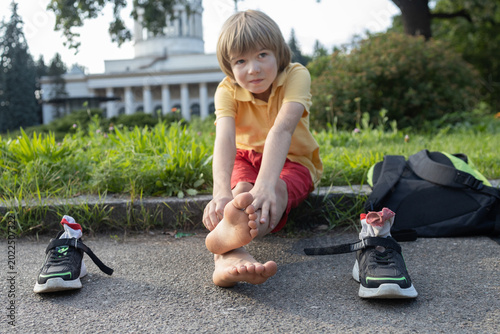 boy sits tiredly on the sidewalk, having removed his sneakers and backpack. Focus on his feet. The child is barefoot and sockless, trying to cool his feet in the hot weather and rest