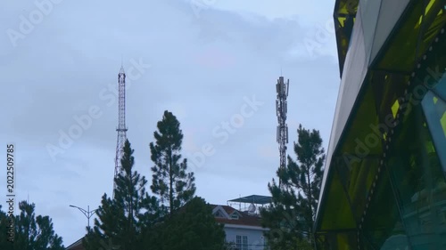 Two telecommunication towers featuring various antenna types typical of wireless infrastructure used for mobile networks rise above pines and other coniferous trees with a glass made building 