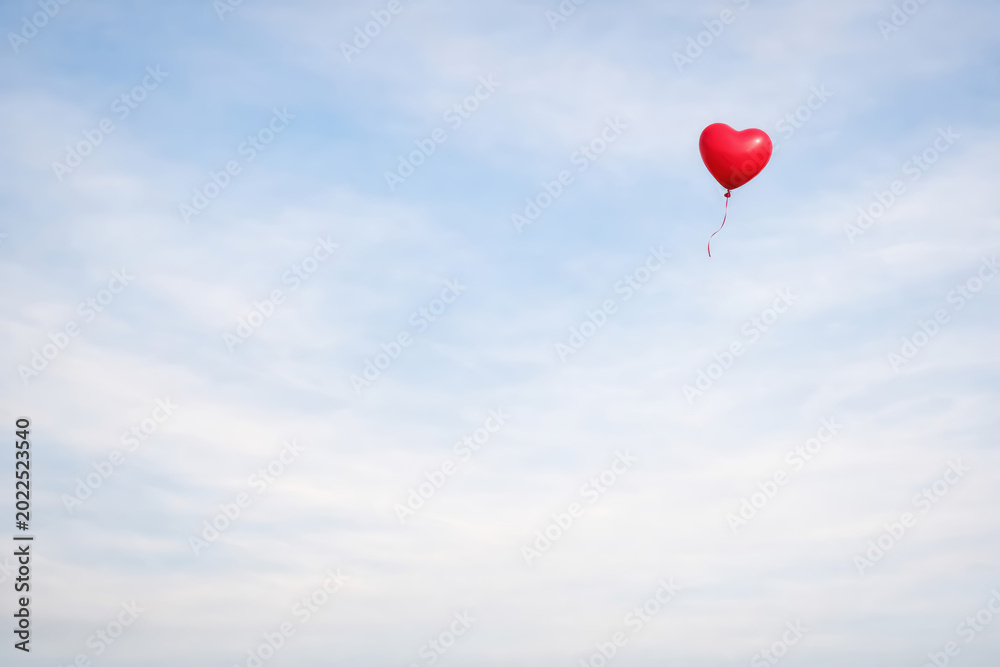Fototapeta premium A single small red heart shaped balloon floating in a bright blue sky with light wispy clouds, representing love, joy, and simplicity