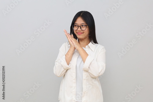 Photos A cunning young Asian woman rubs her hands, planning something with a shy facial expression, glancing sideways into empty space with a smile, standing on a white background