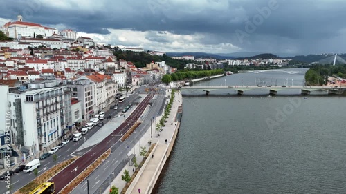 Wide aerial view of Coimbra showing the historic hillside city with riverside boulevard and Mondego River with multiple bridges including a modern pedestrian bridge under dramatic sky
