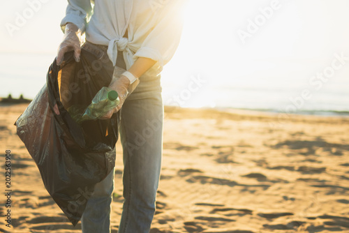 Volunteer collecting plastic waste bottles from a sandy beach at sunset, cleaning up ocean pollution, protecting marine environment, and promoting ecological sustainability