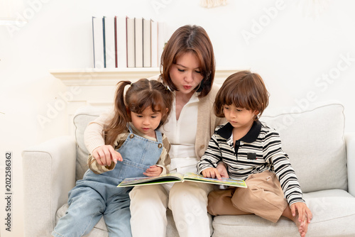 Asian single mother guides daughter, son through page activity, explaining images step by step. Homeschooling learning game strengthens trust, patience, emotional support, family happiness indoors.