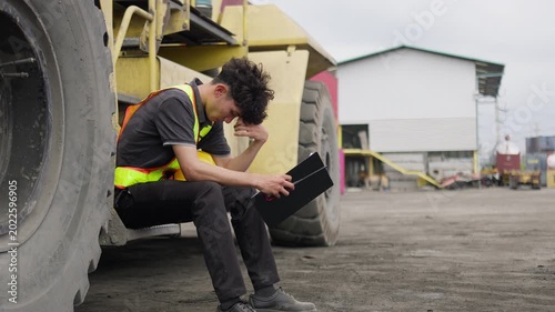 A stressed engineer reviewing a difficult report at an industrial site, sitting on heavy machinery and showing signs of workplace burnout and pressure.