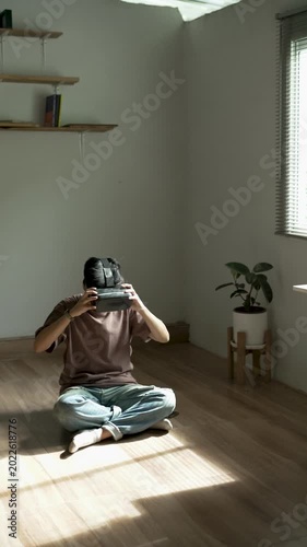 Young man sitting on a wooden floor, putting on a VR headset and adjusting the website to start his journey into the metaverse. Vertical video