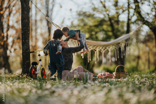 Mother plays with her two young children in a sunlit park while holding the baby above her. A relaxed family picnic scene with a hammock, bike and blanket on grassy spring ground.