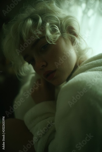 Intimate dreamy close-up portrait of a beautiful young woman with platinum blonde curly hair and freckles, wrapped in a soft white fluffy bathrobe, gentle natural window light, moody pensive expressio