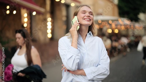Happy young blonde woman talking on smartphone while standing on a busy city street