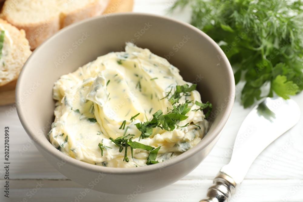Fototapeta premium Delicious butter with herbs in bowl, bread and knife on white wooden table, closeup