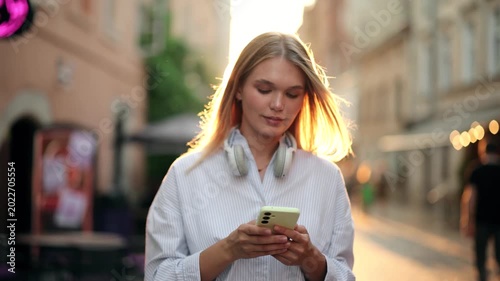 Young blonde woman in headphones texting on smartphone on a city street at sunset